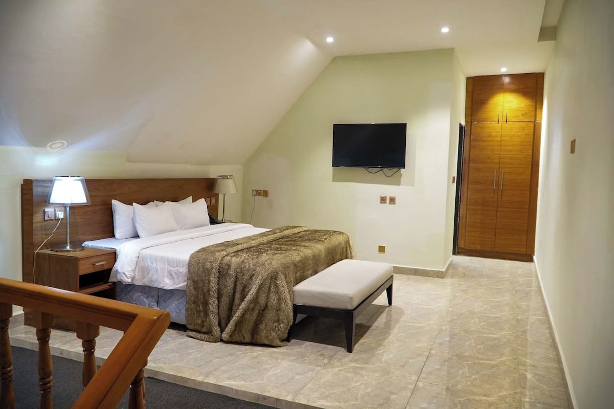 Ein Schlafzimmer mit einem Bett, Nachttisch, Lampe, Bank und einem an der Wand befestigten Fernseher.