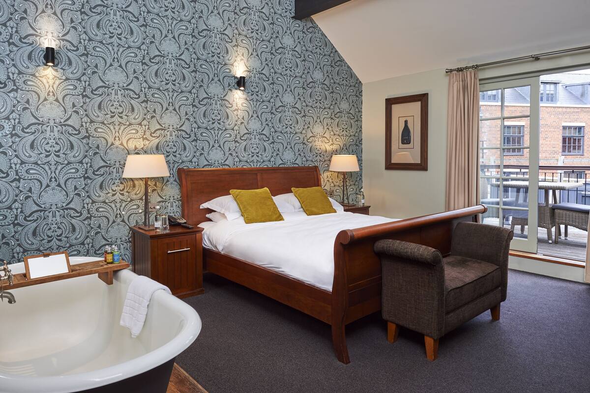 A hotel room with a large bed, two bedside lamps, a wooden nightstand, a large bathtub, and a view of the outside balcony.