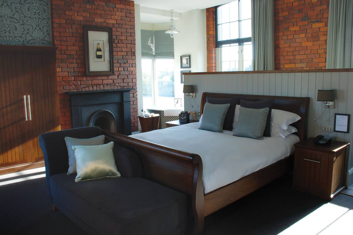 A bedroom with a brick wall, a bed with multiple pillows, a nightstand with a lamp, and a wooden wardrobe.