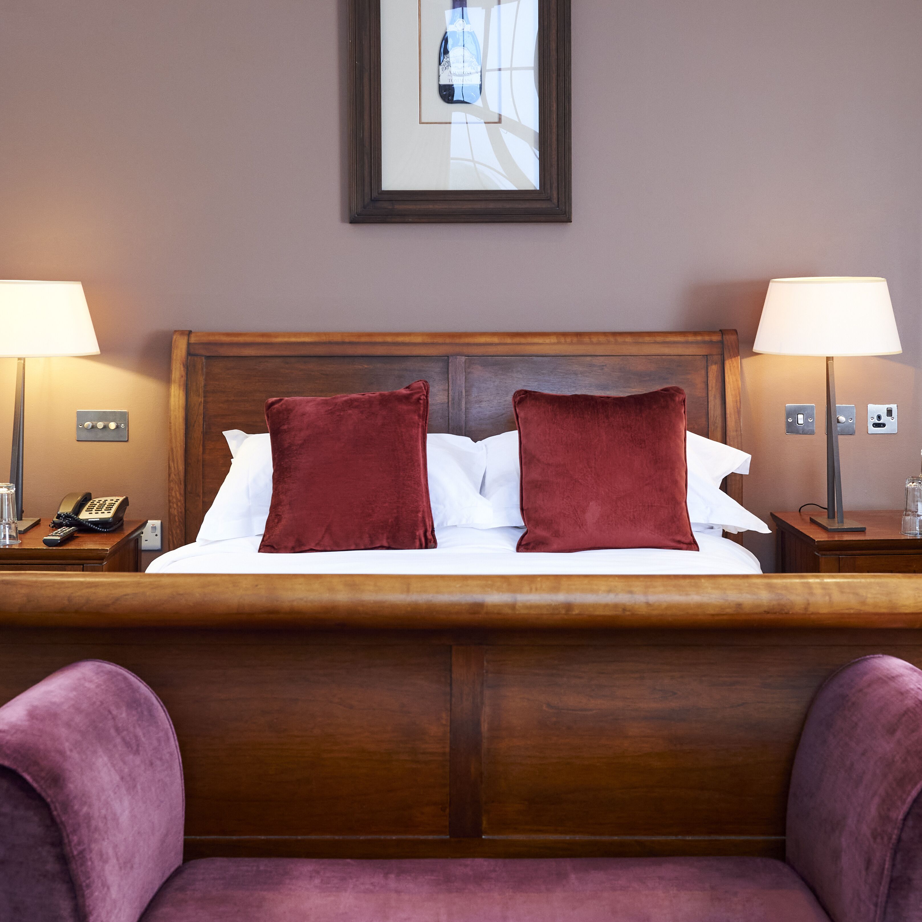 A hotel room with a wooden headboard, two red pillows, white linens, a framed picture on the wall, and two bedside lamps.