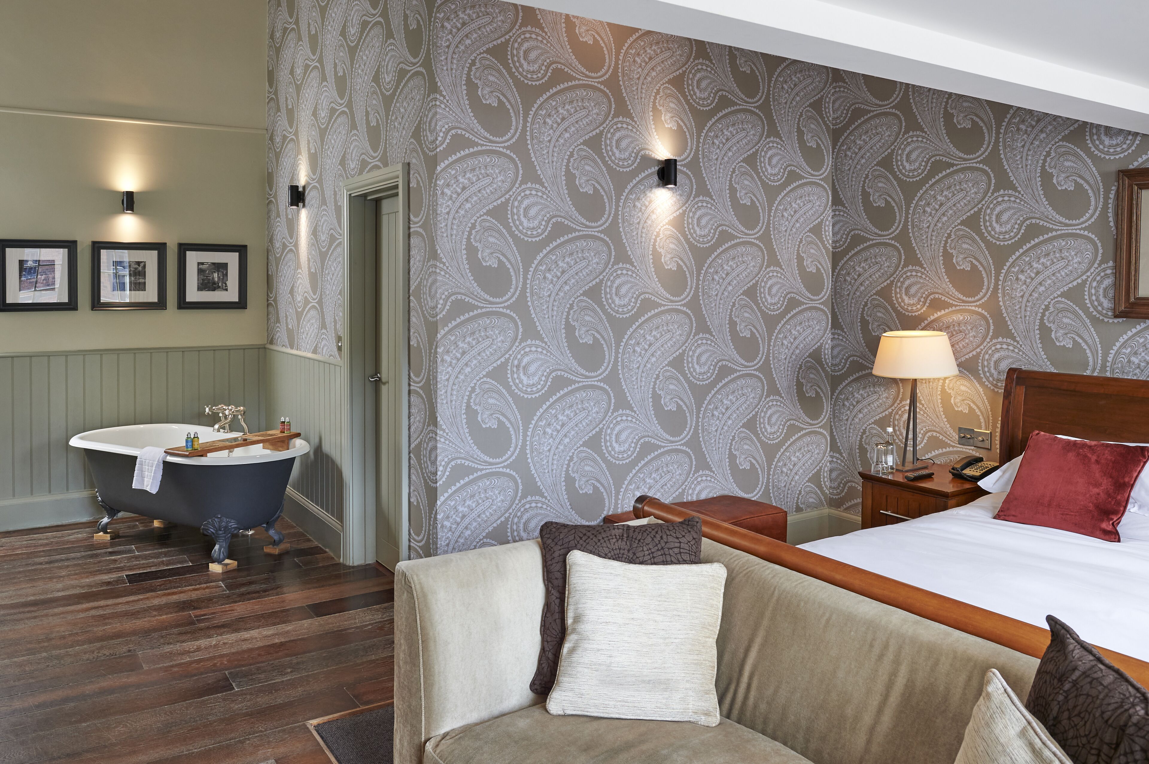 A hotel room with a bathtub, a bed with a red pillow, a wooden nightstand, and a wall with a paisley pattern.