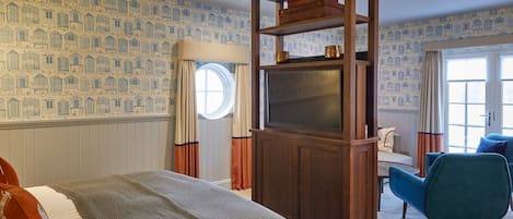 Un dormitorio con una cama grande, un mueble bajo de televisión de madera, un sillón azul y papel tapiz estampado.