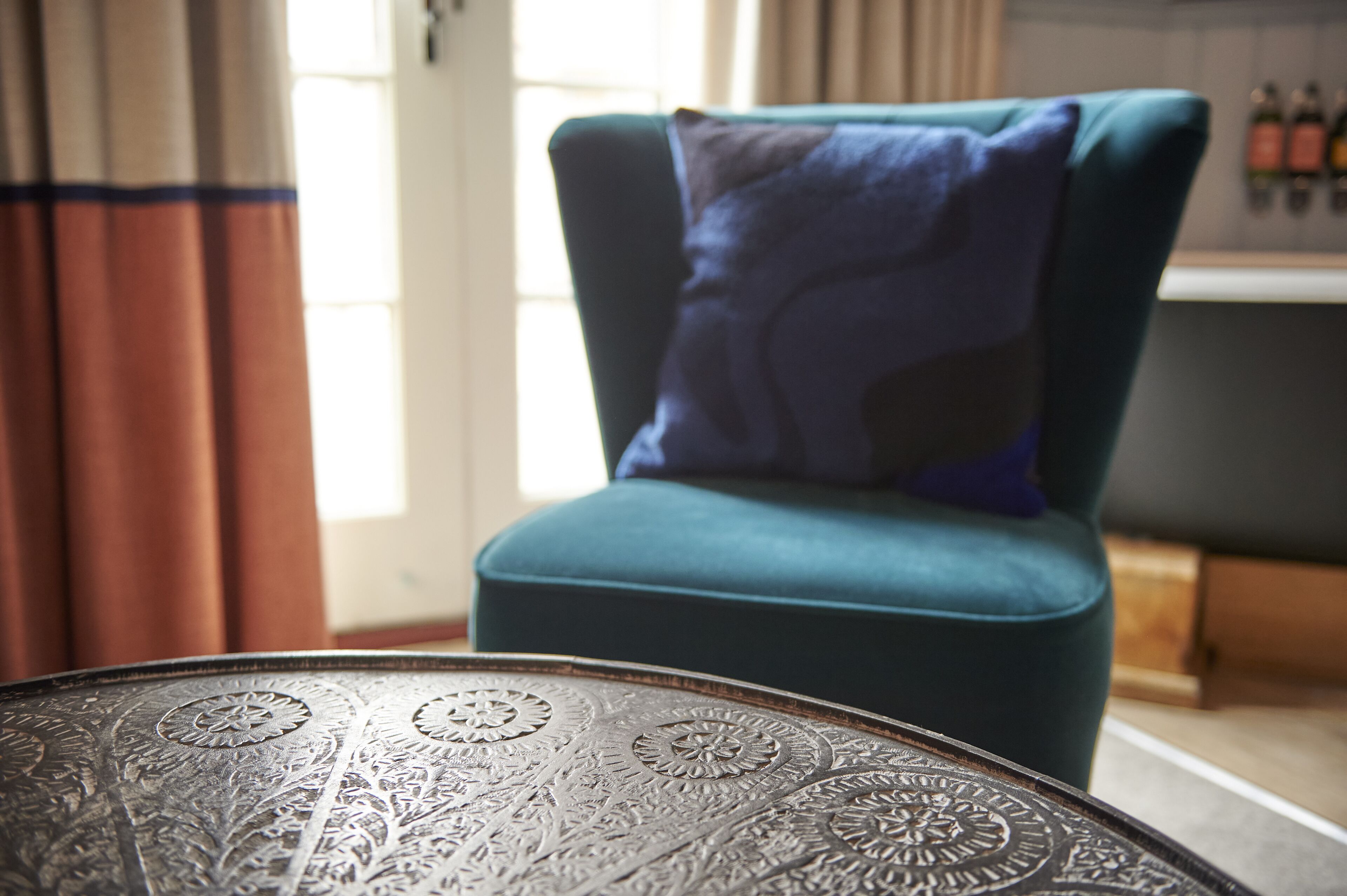 Een teal fauteuil met een blauw kussen, een tafeltje van gestreept metaal en een raam met gordijnen.