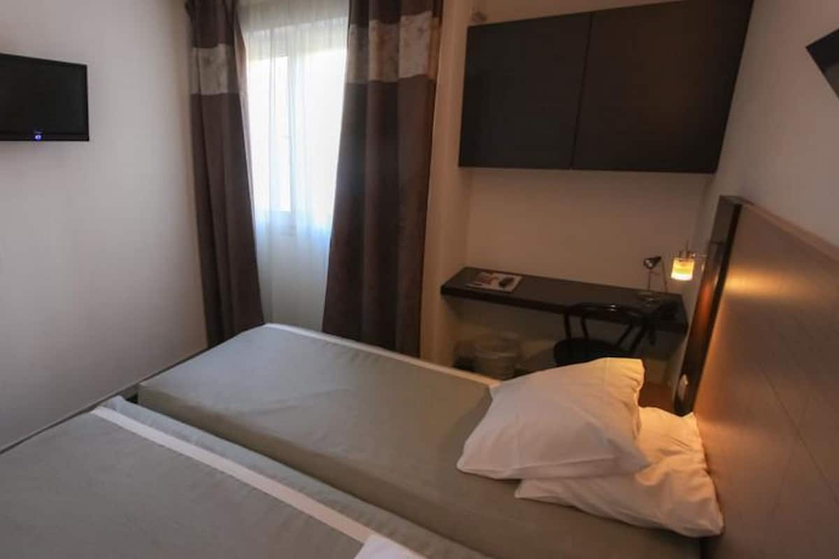 Un lit simple avec un oreiller, un téléviseur fixé au mur, un bureau avec une chaise et une fenêtre avec des rideaux.