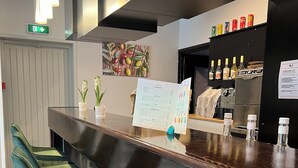 A bar area with a dark counter, green bar stools, a menu stand, and a wall-mounted shelf with bottles. - Adonis Arc Hôtel Aix (Aix-en-Provence)