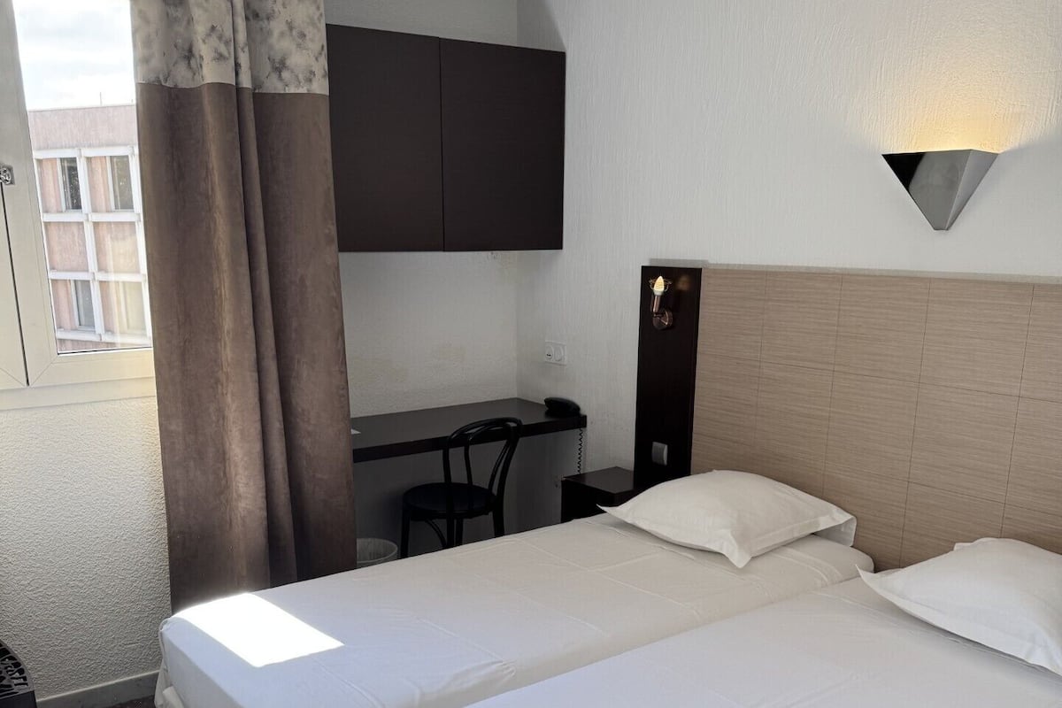 Une chambre d’hôtel avec deux lits, un bureau, une chaise, un téléphone et un luminaire fixé au mur.