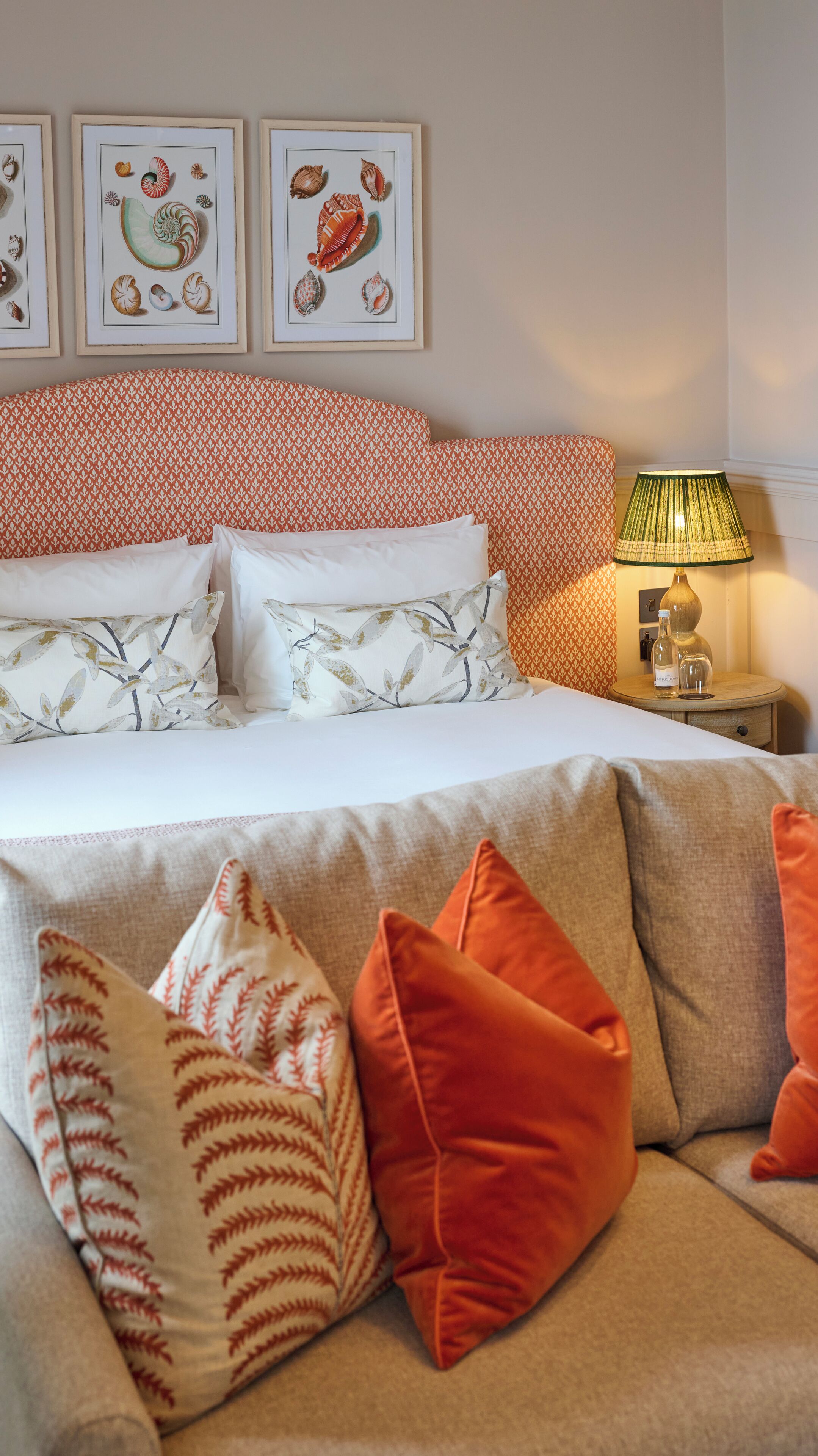 A neatly arranged bedroom with a patterned headboard, multiple pillows, a bedside table with a lamp, and framed artwork on the wall.