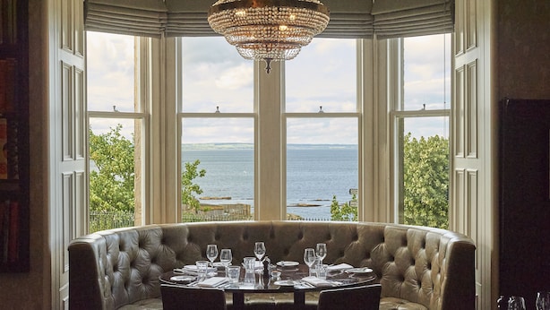 A dining area with a round table set for six, a tufted leather bench, and a view of the sea through large windows.