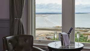 A room with a view of a beach and sea, a table with a bottle and a glass, and a chair.