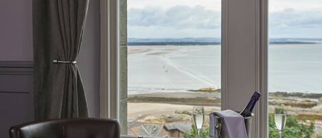 A room with a view of a beach and sea, a table with a bottle and a glass, and a chair.