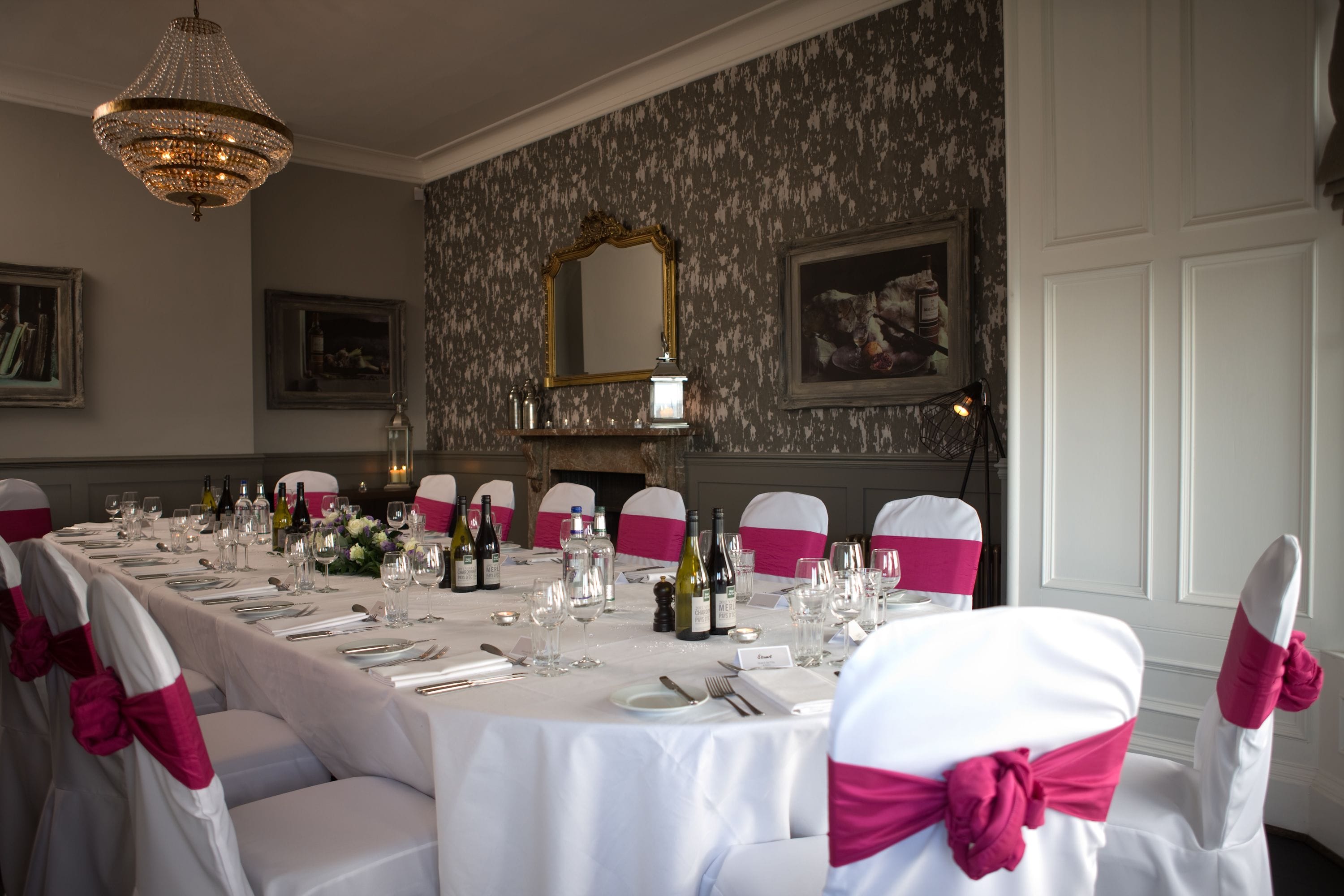 Ein eleganter Speisesaal mit einem langen Tisch, der für eine Mahlzeit gedeckt ist, Stühle mit weißen und rosa Bändern, einer Kandelaber und gerahmten Kunstwerken an den Wänden.