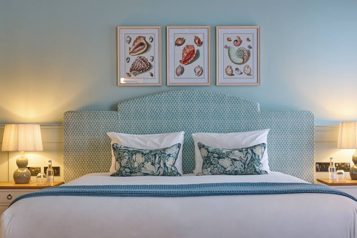Ein ordentlich bezogenes Bett mit Blumenkissen, einem gemusterten Kopfteil und einem Rahmenbild darüber.