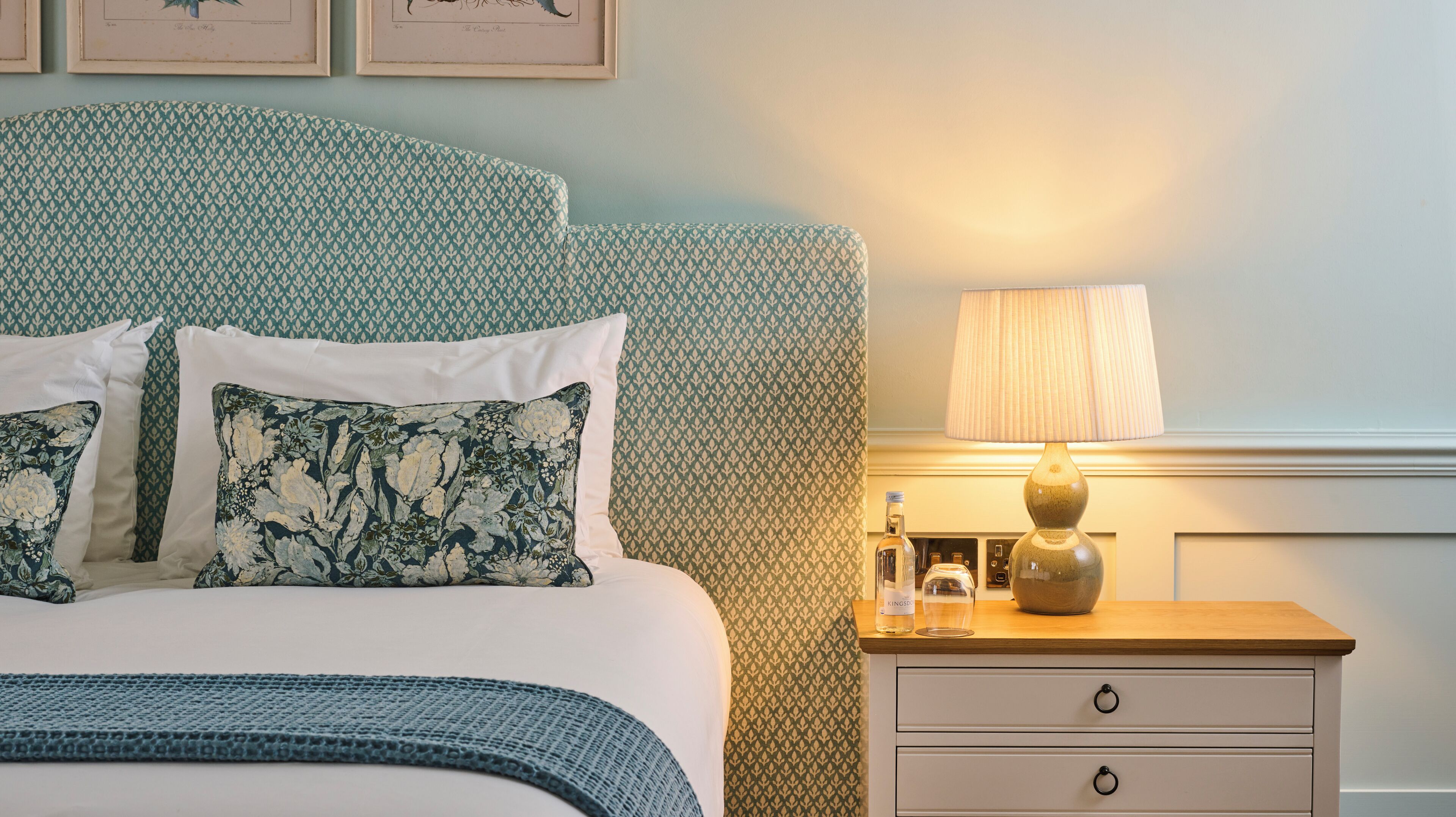 A neatly made bed with floral pillows, a bedside table with a lamp, and a framed botanical illustration on the wall.