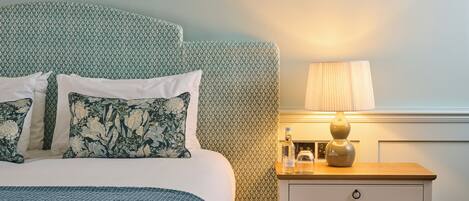 A neatly made bed with floral pillows, a bedside table with a lamp, and a framed botanical illustration on the wall.