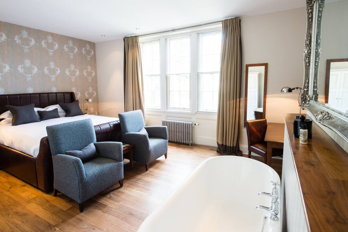 A hotel room with a large bed, two armchairs, a desk, a bathtub, and large windows with curtains.