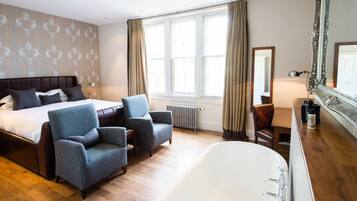 A hotel room with a large bed, two armchairs, a desk, a bathtub, and large windows with curtains.