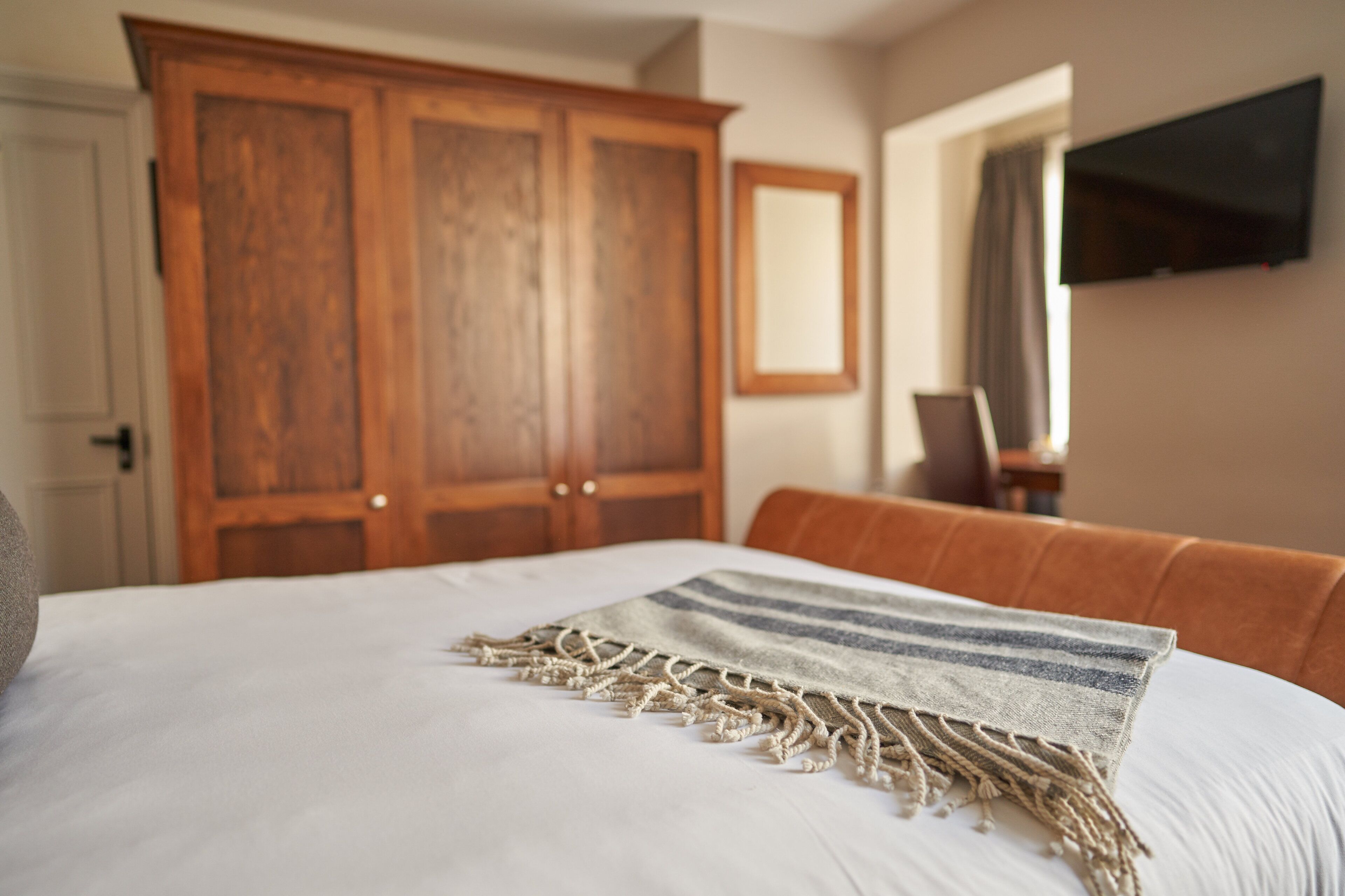 A bedroom with a large bed, a wooden wardrobe, a television mounted on the wall, and a brown leather chair.