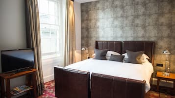 Habitación de hotel con una cama grande, un televisor sobre un soporte de madera, un papel tapiz estampado y una ventana con cortinas.