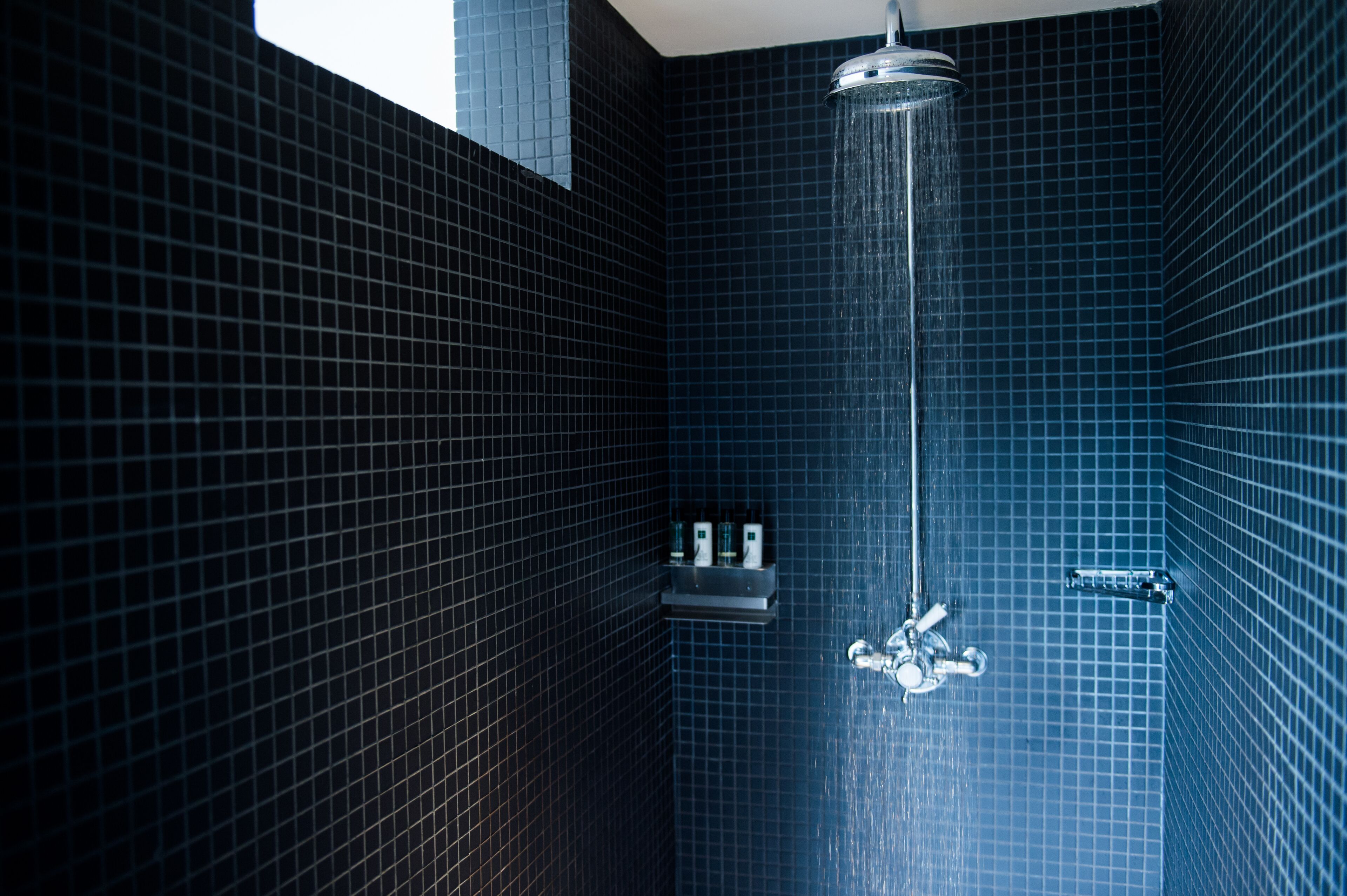 A bathroom with dark blue tiled walls, a shower with a handheld showerhead, and a shelf with toiletries.
