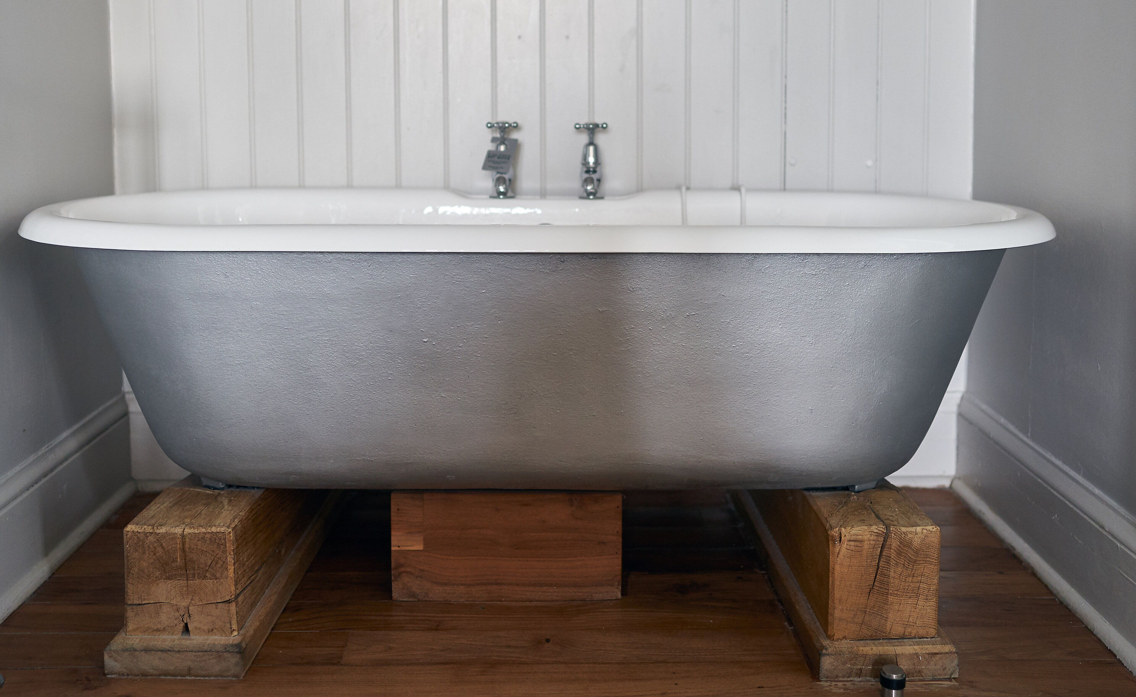 A clawfoot bathtub with a white interior, mounted on wooden supports, in a room with wooden flooring and white paneled walls.