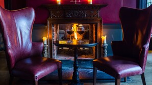 Una habitación con dos sillones de cuero rojo, una mesita redonda con dos copas de vino, una chimenea con velas y un espejo sobre la chimenea.