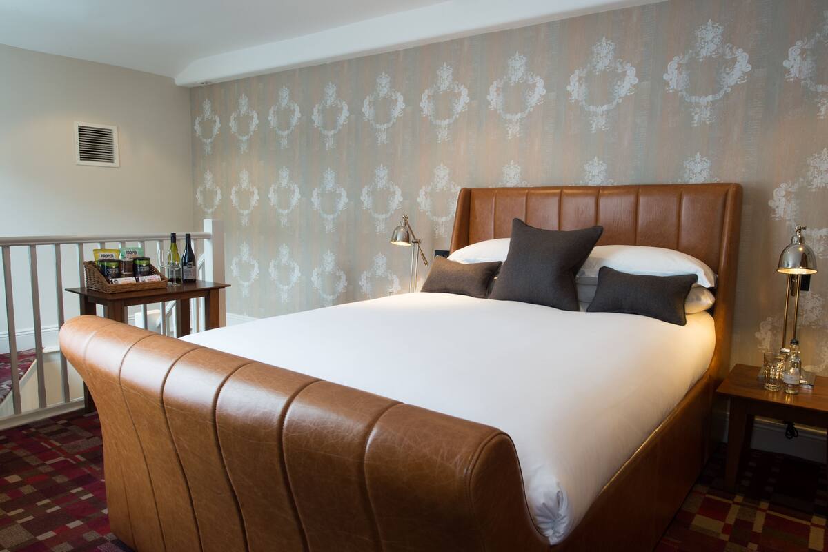 A hotel room with a large bed, a wooden headboard, a nightstand with a lamp, and a small table with a basket of items.