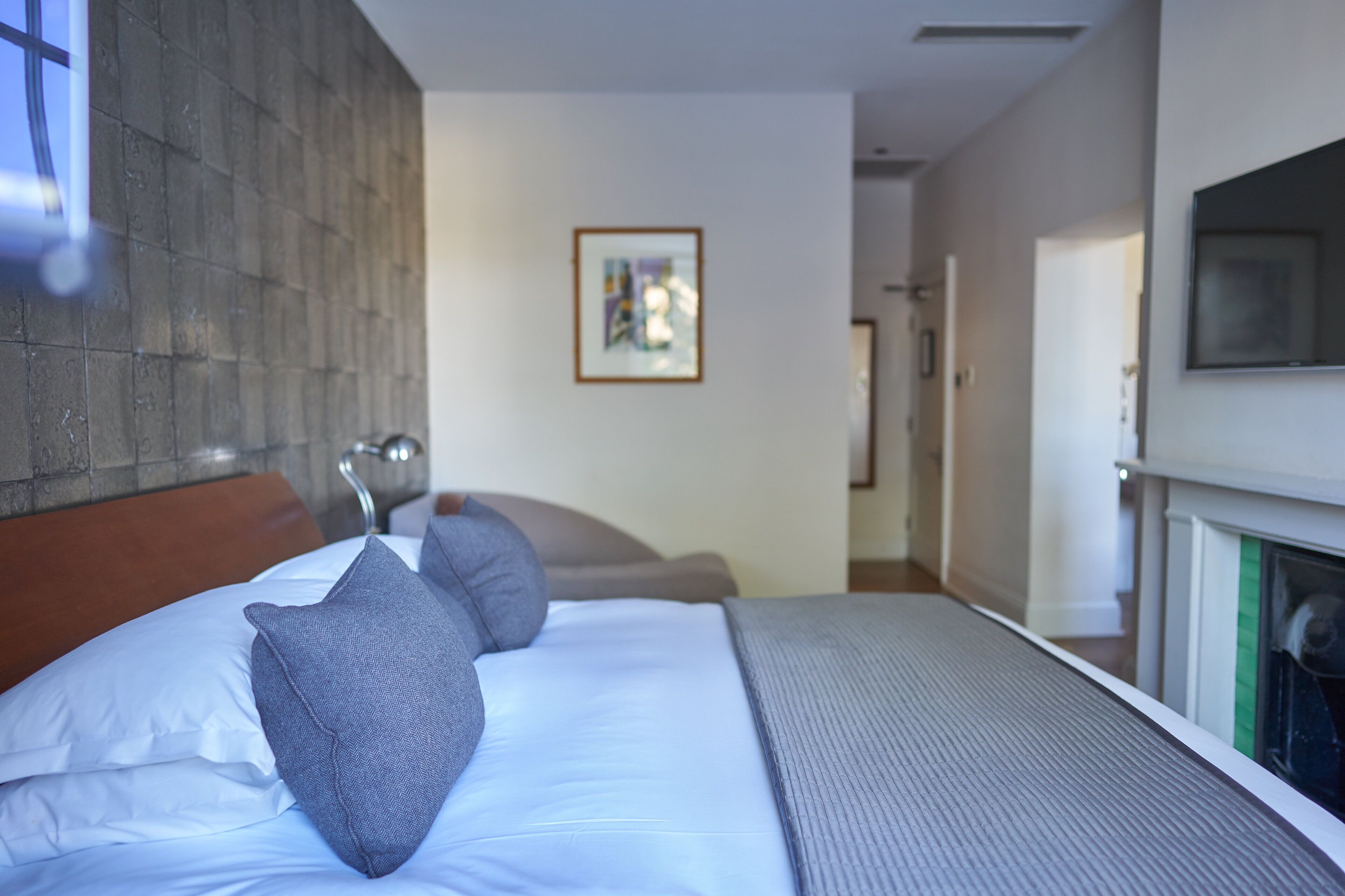 A hotel room with a bed, pillows, a headboard, a painting on the wall, and a fireplace.