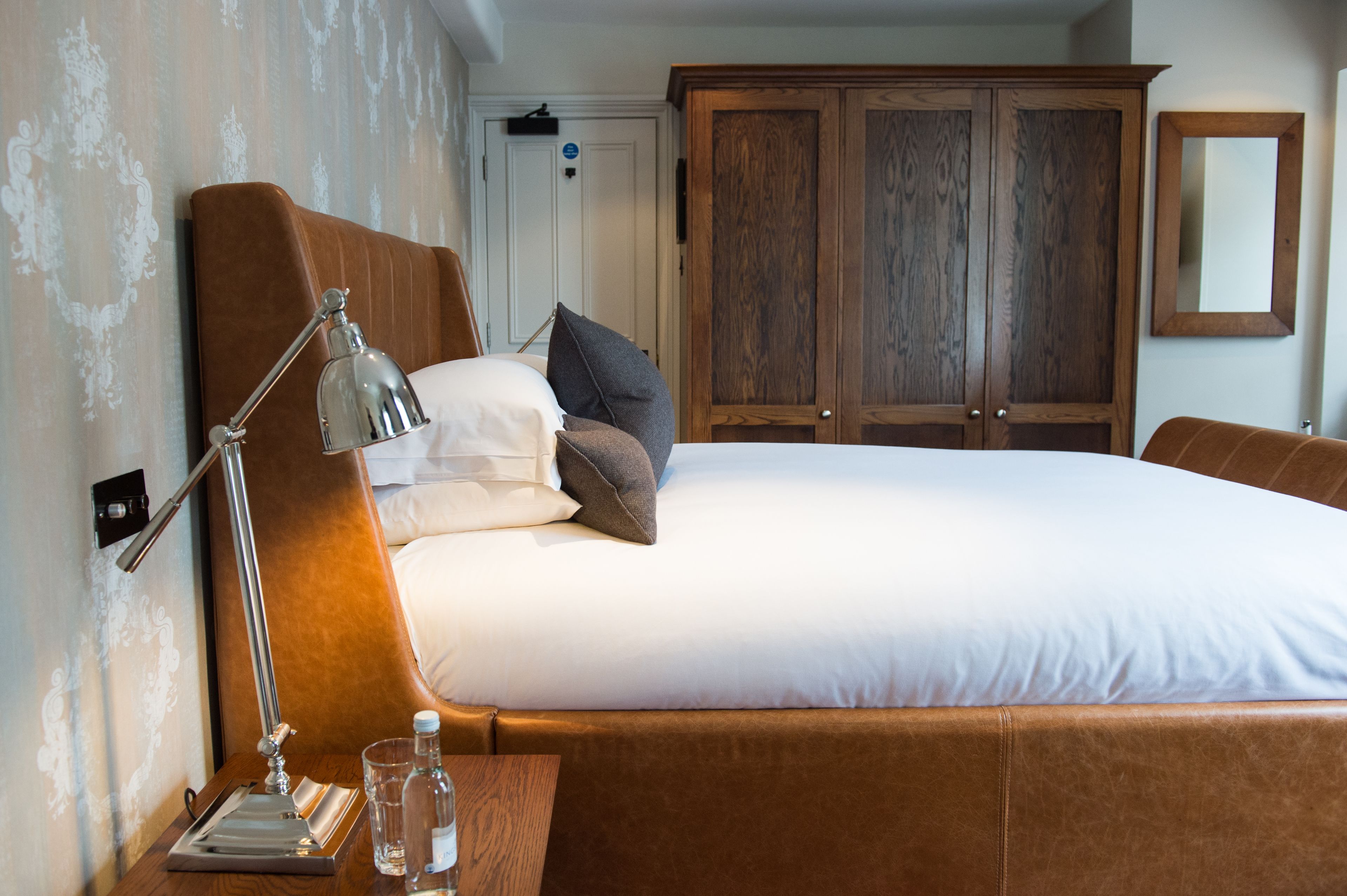 Een net opgemaakt bed met kussens, een bedlampje en een nachtkastje met glazen en een fles.