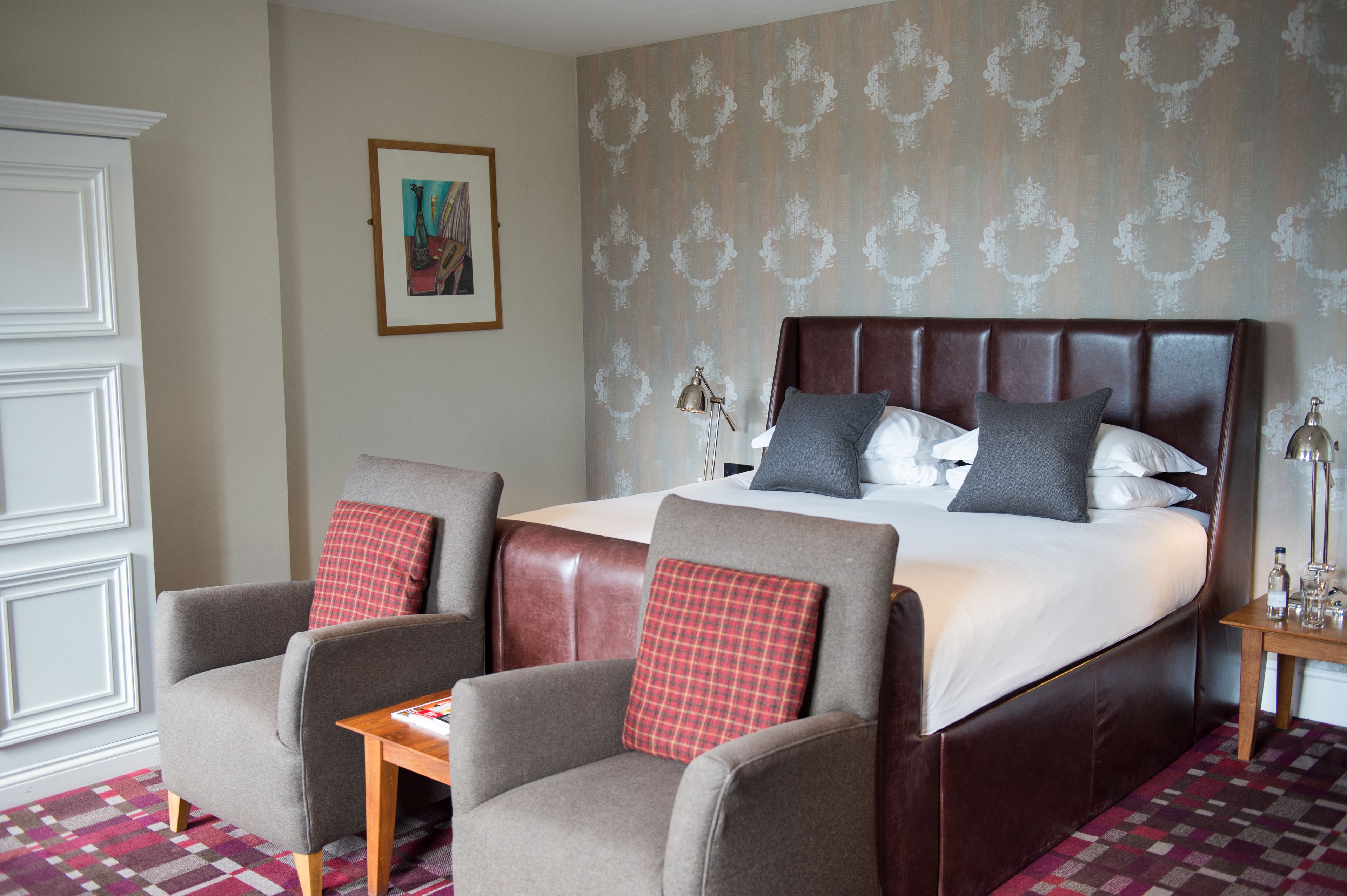 A hotel room with a large bed, two armchairs, a small table, and a nightstand with a lamp.