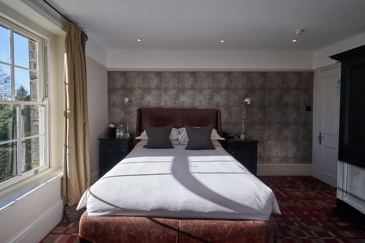 A hotel room with a large bed, two bedside tables, a nightstand, a window with curtains, and a patterned wall.