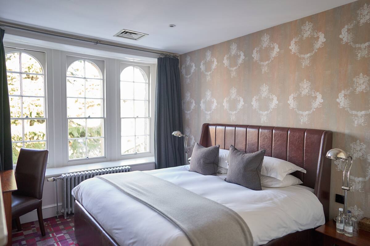 A neatly made bed with a headboard, a chair, a bedside table with a lamp, and a window with curtains.