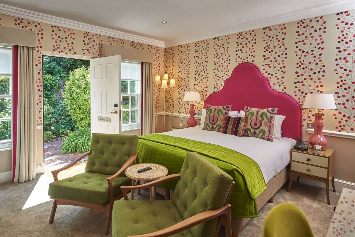 Un dormitorio con cama, sillones tapizados en verde, una mesita de noche con lámpara y vistas al exterior con vegetación.
