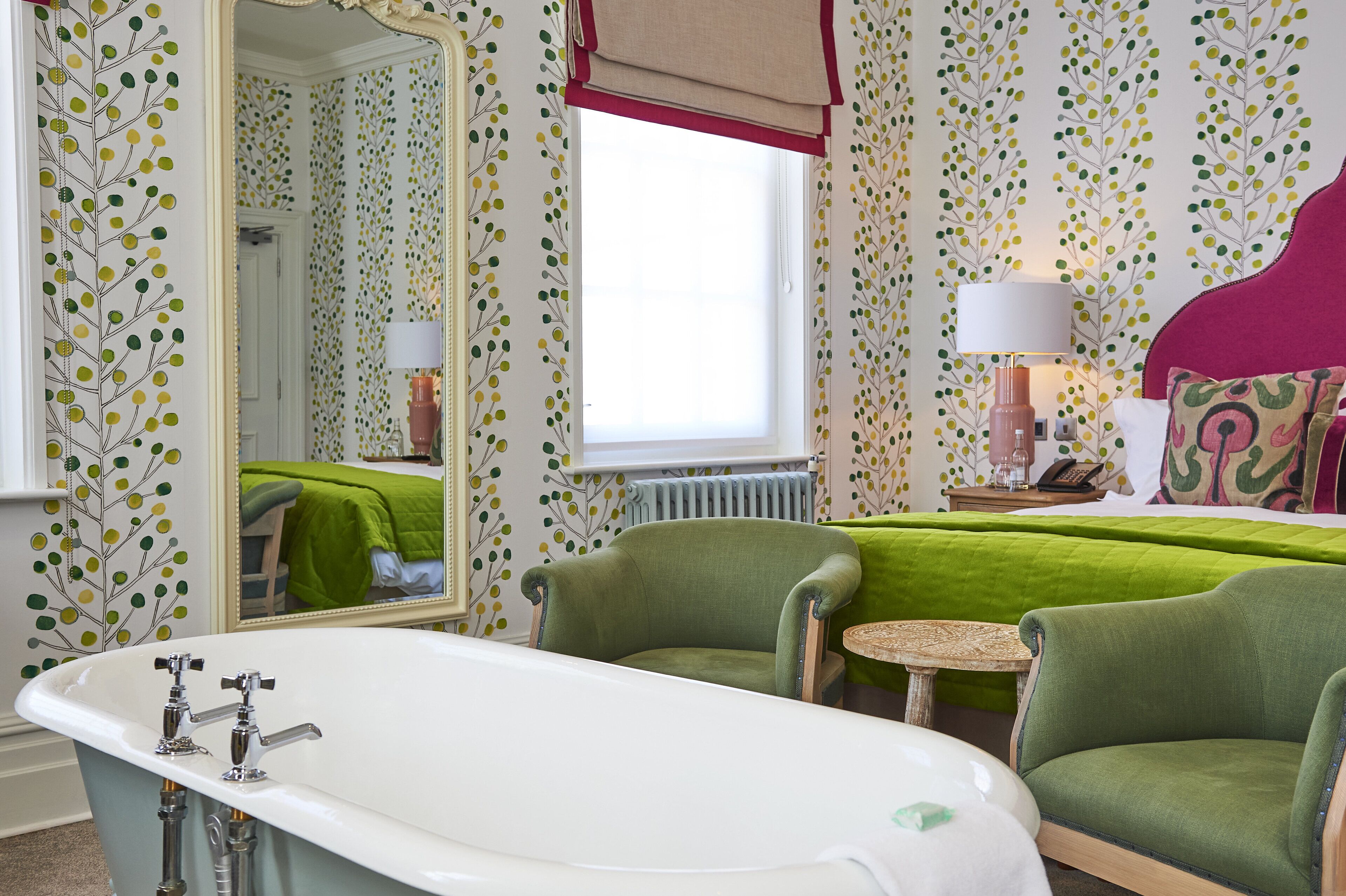 A room with a bathtub, two green armchairs, a small round table, a bed with a green bedspread, a mirror, and a window with a red valance.