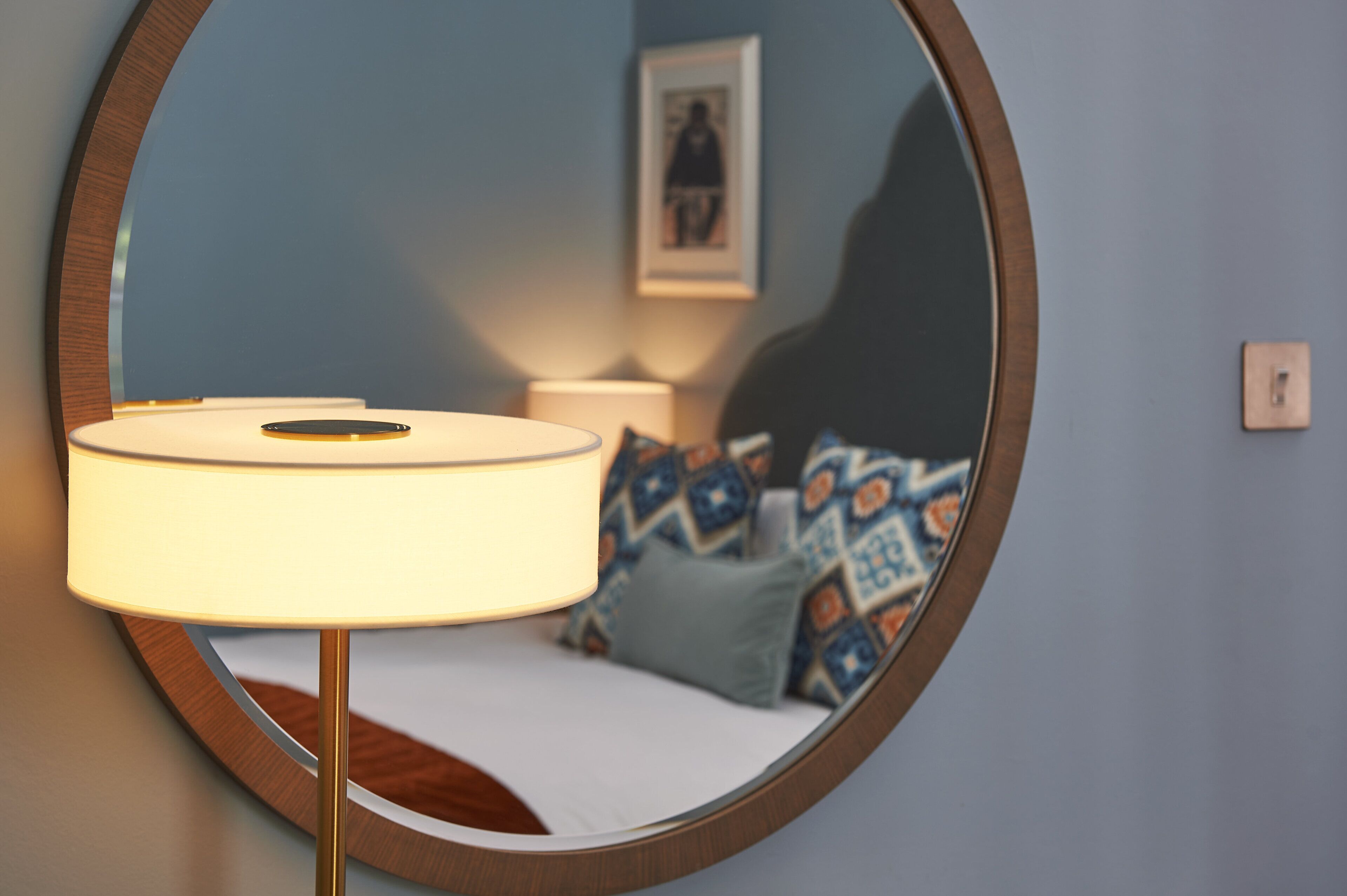 A round mirror reflecting a bed with patterned pillows, a bedside lamp, and a framed picture on the wall.