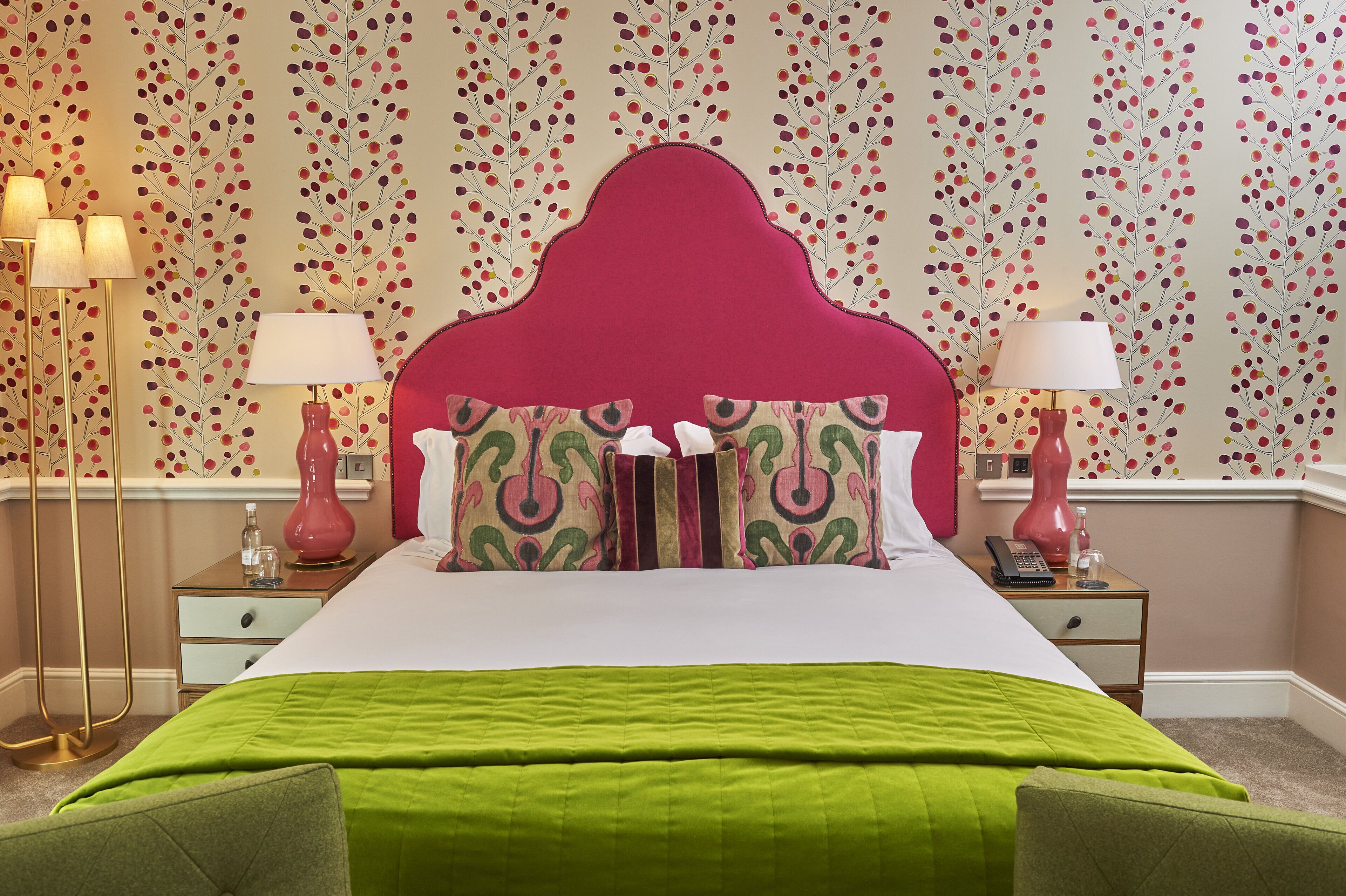 Cama bem arrumada com colcha verde, cabeceira vermelha e almofadas estampadas. Duas mesinhas de cabeceira com luminárias e um pequeno relógio. A parede possui um papel de parede com padrão floral.