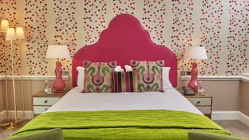 Cama bem arrumada com colcha verde, cabeceira vermelha e almofadas estampadas. Duas mesinhas de cabeceira com luminárias e um pequeno relógio. A parede possui um papel de parede com padrão floral.