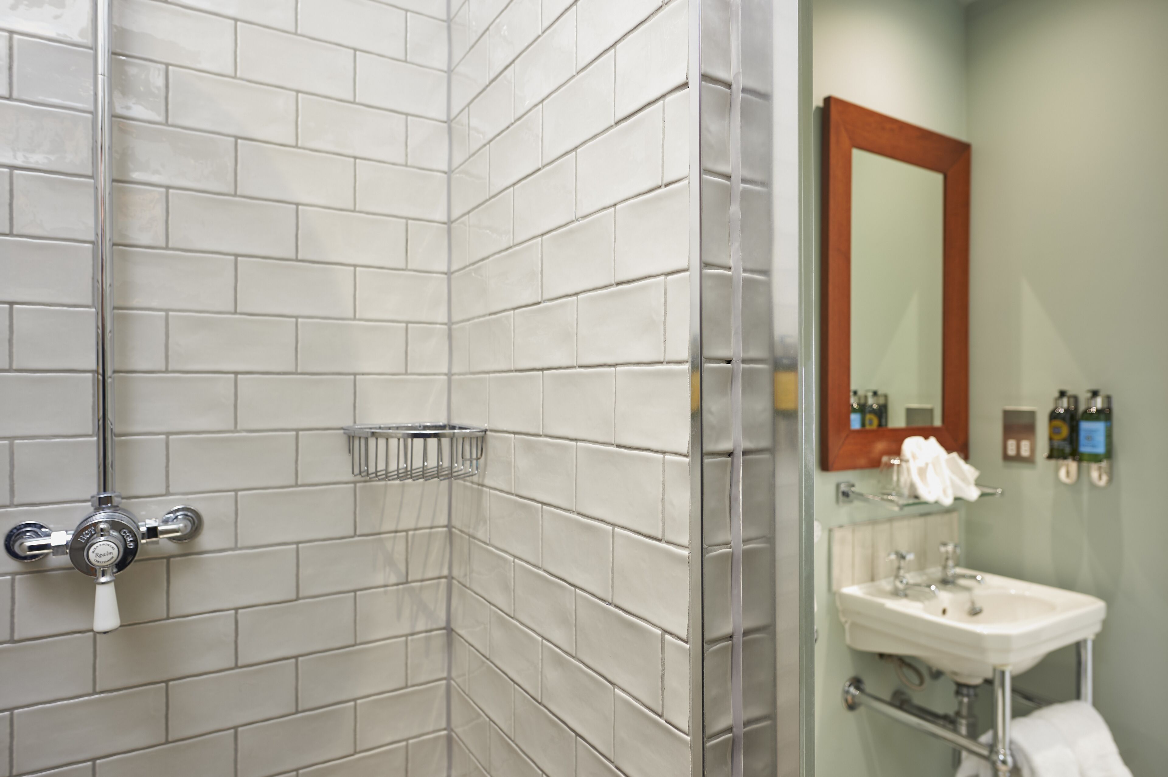 A bathroom with white tiled walls, a shower with a chrome faucet, a glass shelf, and a sink with a mirror above it.