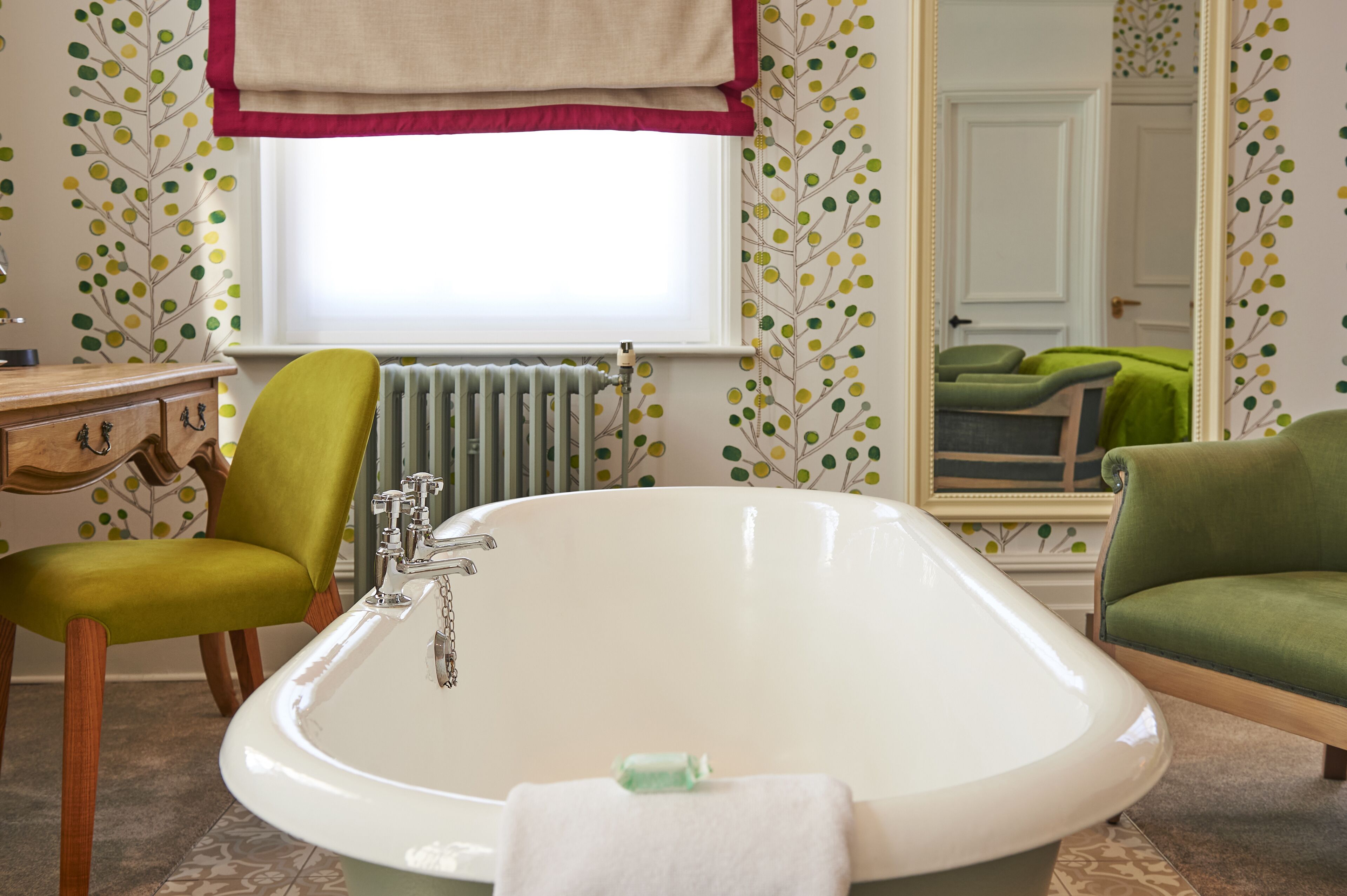 A bathroom with a white bathtub, a green chair, a wooden vanity, and a radiator.
