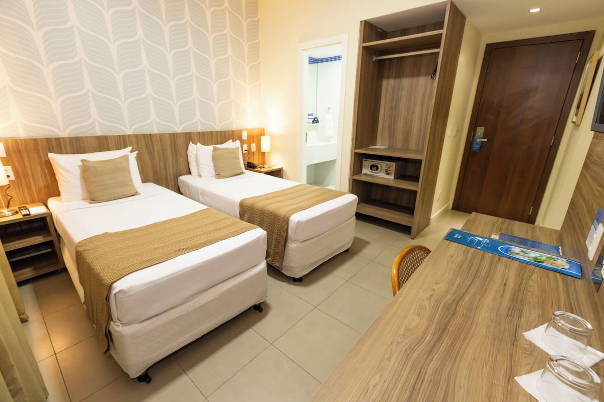 A hotel room with two beds, a desk, a chair, a wardrobe, and a bathroom visible through an open door.