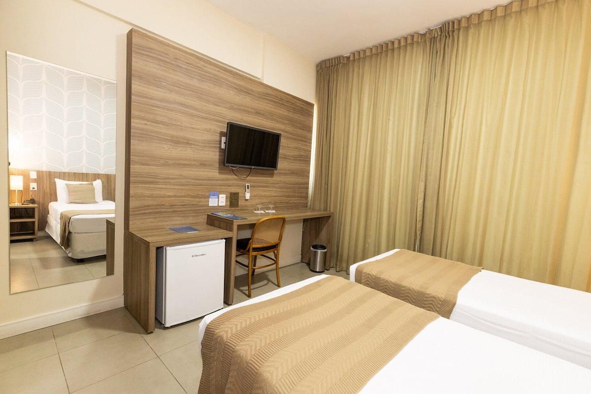A hotel room with a bed, a desk with a computer, a TV, and a small fridge.