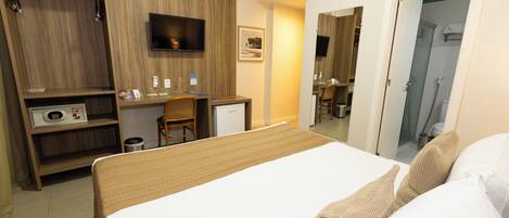 Quarto de hotel com uma cama, escrivaninha, cadeira, televisão e guarda-roupa.