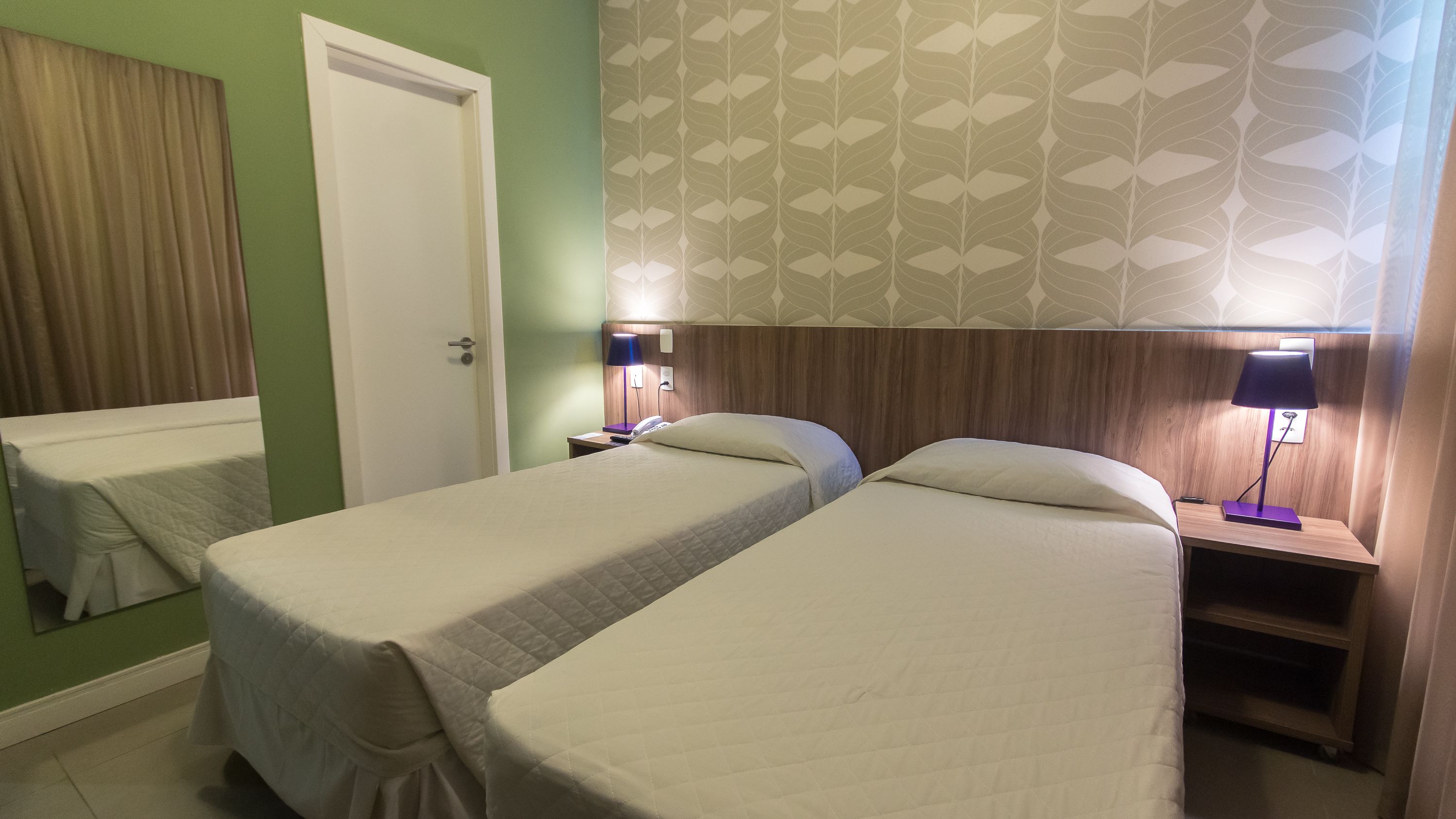 A hotel room with two single beds, a wooden headboard, a nightstand with a lamp, and a green wall.