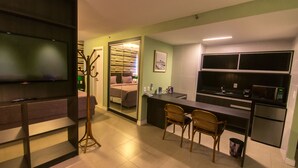 A compact hotel room with a bed, a desk, a chair, a television, a mirror, and a kitchenette. - Mais Hotel Aeroporto Salvador (Lauro de Freitas)