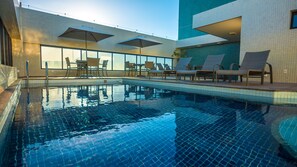 An indoor swimming pool with tiled walls, lounge chairs, and umbrellas.