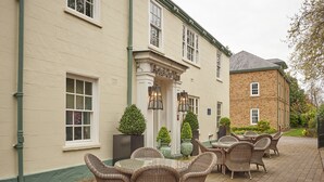 L’extérieur d’un hôtel avec une terrasse dallée, des meubles en osier et un bâtiment en briques.