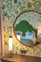 A room with a green headboard, floral wallpaper, a round mirror, and a bedside table with a lamp.