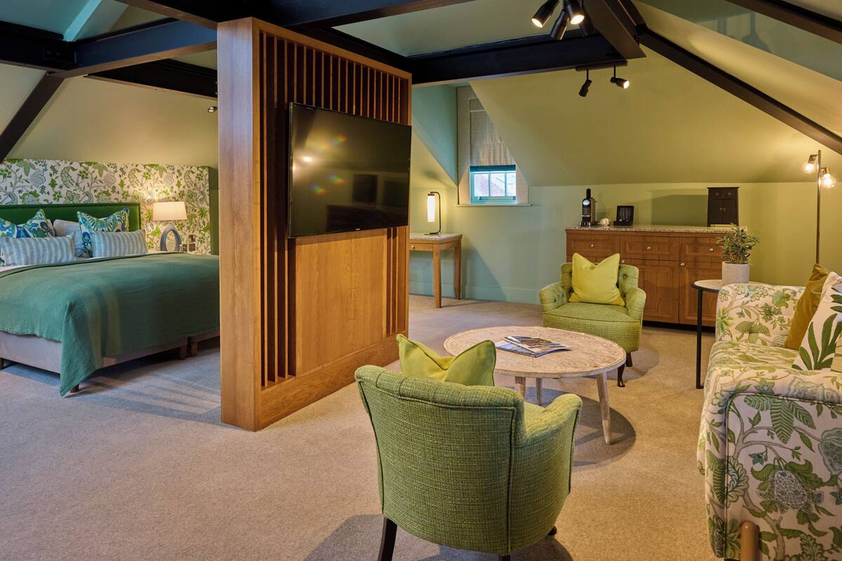 A bedroom with a bed, a TV, and a nightstand. There is a living area with a sofa, a coffee table, and a lamp. The room has wooden beams on the ceiling and a window with a view of the outside.
