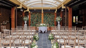 Un lieu de réception pour mariages avec des rangées de chaises en bois, une allée centrale agrémentée de décorations florales et un mur en briques en arrière-plan.