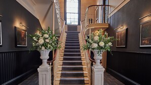 Un magnifique escalier recouvert d’une moquette sombre, flanqué de colonnes blanches ornées de compositions florales, menant à un palier doté d’une grande fenêtre.