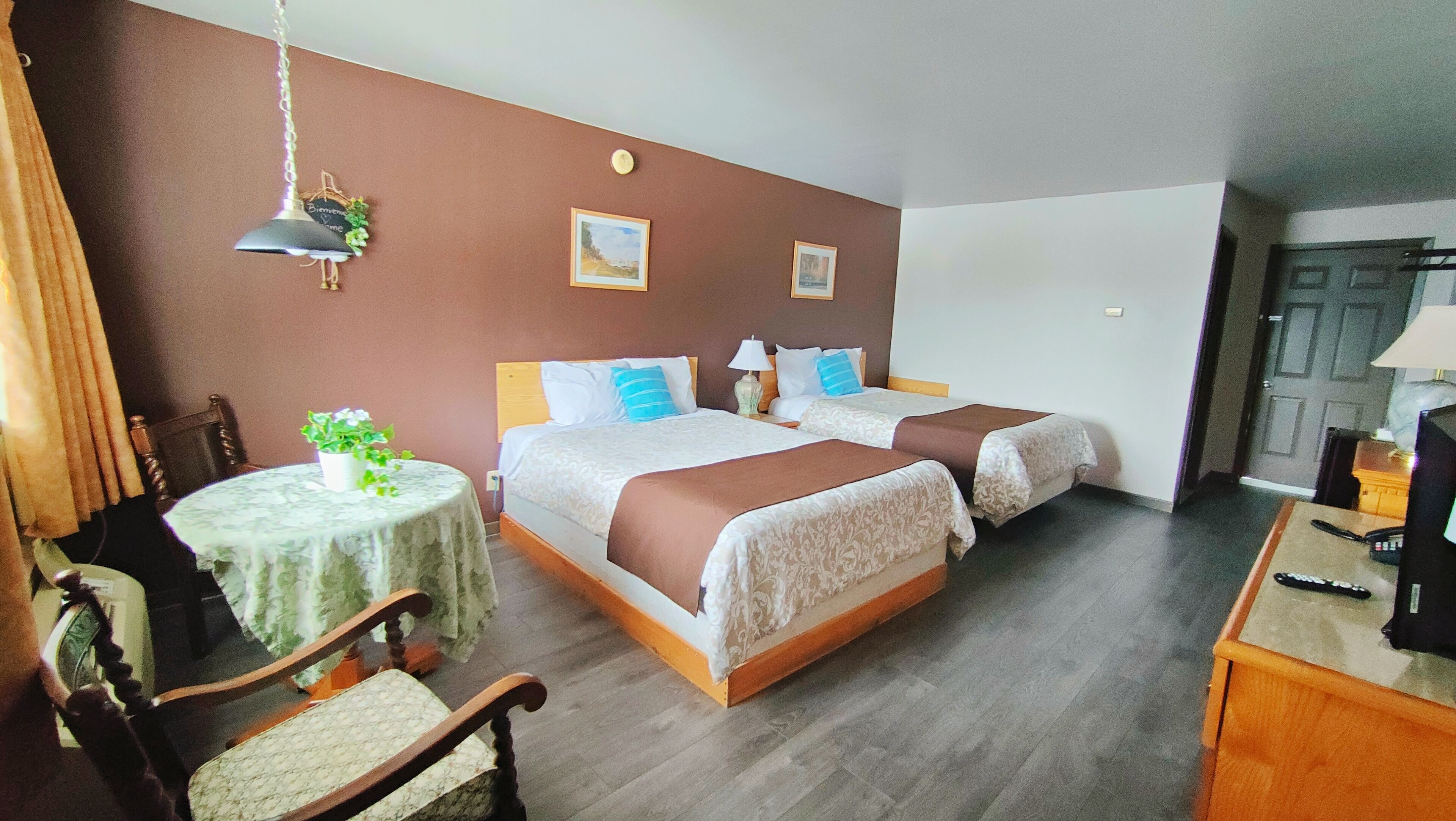 A hotel room with two beds, a small round table with a plant, a chair, a desk with a lamp, and a door.